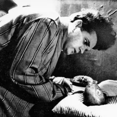 Młody David Lynch w pasiastym szlafroku pochyla się nad stołem, na którym leżą dziwne przedmioty.