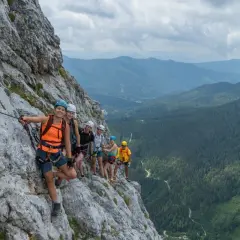Grupa uśmiechniętych ludzi na via ferrata w Dolomitach, zabezpieczona linami, podziwia malownicze widoki górskiego krajobrazu.