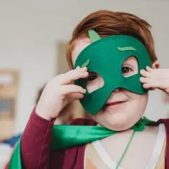 Ein Kind mit roten Haaren trägt eine grüne Drachenmaske und einen Umhang. Das Kind schaut neugierig durch die Augenlöcher.