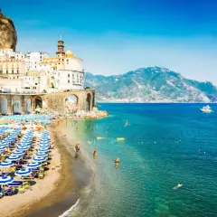 A vibrant beach scene in Naples, with rows of blue umbrellas and sunbathers. Buildings cling to a cliffside, overlooking the sparkling turquoise sea.