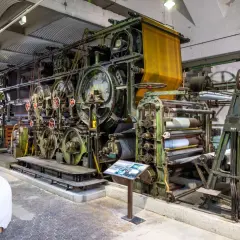 Große, grüne Textilmaschine im lvr industriemuseum, die Stoffbahnen verarbeitet. Eine gelbe Stoffbahn hängt oben, daneben Papierrollen.
