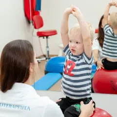 Fizjoterapeutka wspiera chłopca z zaburzeniami rozwojowymi podczas ćwiczeń na piłce rehabilitacyjnej.