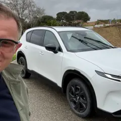 Mężczyzna w okularach pozuje do selfie obok białej Mazdy CX-5 na tle winnicy.