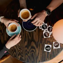 Dwie osoby piją kawę przy drewnianym stoliku. Czy kawa kuloodporna przerywa post? Na stoliku leżą też naklejki z napisem "GOOD COFFEE IS A HUMAN RIGHT".