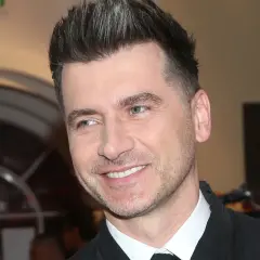 Uśmiechnięty Tomasz Kammel, jego wiek nie jest widoczny, ale jego stylizacja i fryzura sugerują, że jest w sile wieku.