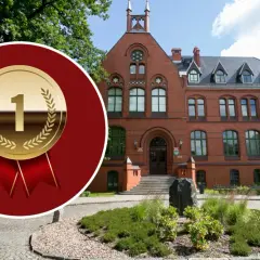 Złoty medal z cyfrą 1 na tle ceglanego budynku i zieleni.