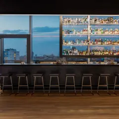 Interni di un locale con bancone bar, sgabelli e scaffali pieni di bottiglie. Vista sulla città al tramonto, un'atmosfera da next club Torino.