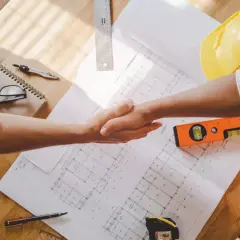 Uścisk dłoni nad planami budowlanymi, obok kasków i poziomicy. To symbol finalizacji prostej umowy o roboty budowlane - wzór.