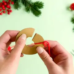 Hände wickeln roten Faden um Pappring für **Basteln mit Pompons Weihnachten**. Tannenzweige und Kugeln im Hintergrund.