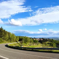 Strada SR 222 Chiantigiana serpeggia tra colline verdi e boschi, con un borgo toscano in lontananza sotto un cielo azzurro e nuvoloso.