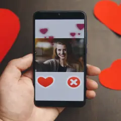 Dłoń trzyma smartfon z ekranem aplikacji randkowej. Widoczna uśmiechnięta dziewczyna i przyciski serca i krzyżyka. Wokół czerwone serca.