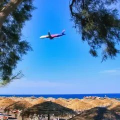 Samolot przelatuje nad plażą z parasolami, tuż przed lądowaniem na **Santorini lotnisko**.