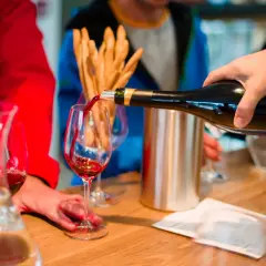Ręka nalewa czerwone wino do kieliszka. W tle widać ludzi i przekąski.