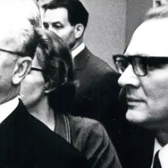 Ein Mann mit Brille und Bart, ein DDR-Chef, lächelt. Im Hintergrund weitere Personen.