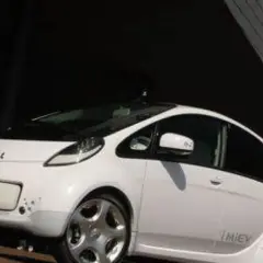 Biały, kompaktowy Mitsubishi i-MiEV stoi przed nowoczesnym budynkiem.