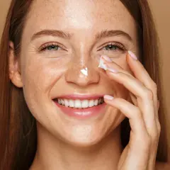 Uśmiechnięta kobieta z piegami aplikuje krem na nos. Pamiętaj o ochronie przed słońcem, zwłaszcza stosując retinol.