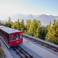 Czerwony wagon kolejki zębatej jedzie po torach w górę zbocza. W tle góry i lasy.