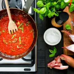 En kastrull med bubblande tomatsås och en träsked. Ingredienser för ett tomatsås recept: färska tomater, schalottenlök, basilika och salt.