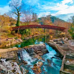 Cascada y puente de madera en la vía verde Puente Viesgo, rodeados de árboles otoñales y un cielo azul vibrante.