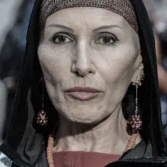 Kobieta w kapturze i ozdobnej opasce na głowie. Jej twarz, naznaczona kora wiek, opowiada historię. Długie kolczyki i naszyjnik dodają jej charakteru.