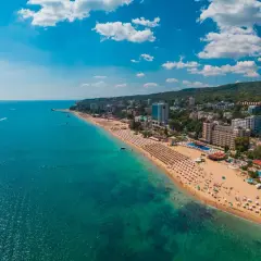 Słoneczna plaża z mnóstwem leżaków i parasoli, tętniące życiem kurorty nad morzem, a w oddali zielone wzgórza. Idealne miejsce na wakacje.