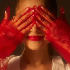 Ariana Grande 2024 w czerwonych rękawiczkach zakrywa oczy, odsłaniając usta i tatuaże na dłoniach.