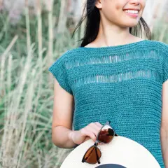 Donna sorridente con un top uncinetto blu petrolio, tiene un cappello di paglia e occhiali da sole.