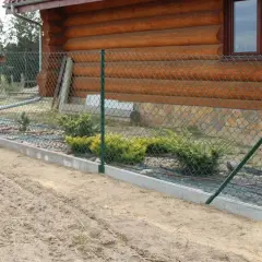 Zielona siatka ogrodzeniowa, zamontowana na betonowym podmurówce, otacza drewniany dom. Wokół rosną krzewy.