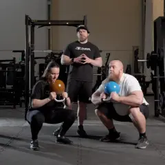 Två personer utför goblet squat med kettlebells medan en instruktör står bakom.