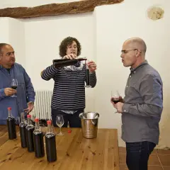 Weinprobe: Eine Frau gießt Wein ein, während zwei Männer zuschauen. Flaschen und Gläser stehen bereit für die assemblage wein.