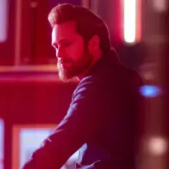 Mężczyzna z brodą w ciemnej koszuli odwraca się od neonowego napisu. Scena z programu telewizyjnego "Bastion".