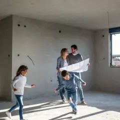 Rodzina ogląda plany w mieszkaniu w stanie deweloperskim. Dzieci biegają po pustym pokoju, rodzice analizują projekt.