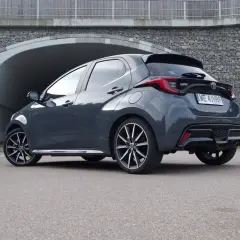 Szary Toyota Yaris GR Sport z czarnym spojlerem i dyfuzorem, zaparkowany przed tunelem z kamiennym murem.