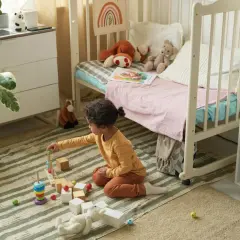 Ein kleines Kind spielt mit Holzbausteinen in einem gemütlichen, quadratischen Kinderzimmer. Spielzeug und ein Bett laden zum Verweilen ein.