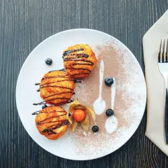 Drei goldbraune, knusprige Kugeln, beträufelt mit Schokoladensauce, liegen auf einem Teller mit Kakaopulver und Beeren. Ein süßer Genuss, der an Eis erinnert, aber als knusprige Leckerei serviert wird.