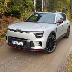 En smart 5-dörrars SUV i silver med röda detaljer kör på en grusväg genom en höstskog.