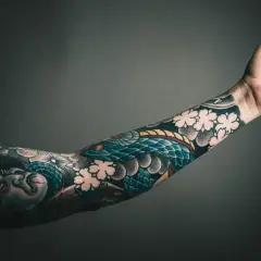 Ein Arm mit einem detailreichen Tattoo, das eine Schlange und Kirschblüten zeigt. Die Botschaft "alles wird gut" ist in diesem Kunstwerk auf der Haut verewigt.