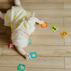 Niemowlę w żółtej sukience w kropki bawi się na podłodze, próbując obrócić się z brzucha na plecy. Wokół leżą kolorowe puzzle ze zwierzątkami.