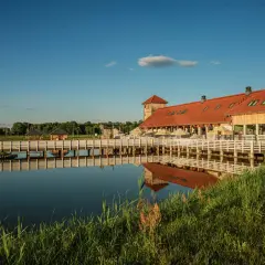 Drewniany kompleks wypoczynkowy nad jeziorem, z pomostem i altanami. W oddali widać wieżę i budynek z czerwoną ruda sułowska.