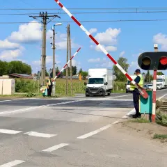 Przejazd kolejowy z zaporami, sygnalizacją świetlną i pracownikami obsługi. Samochody czekają na przejazd.