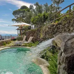 Piscina con cascata che ricorda le terme naturali di Ischia, circondata da vegetazione e con vista sul mare.