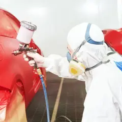 Pracownik w kombinezonie ochronnym wykonuje lakierowanie całego samochodu na czerwono za pomocą pistoletu natryskowego.