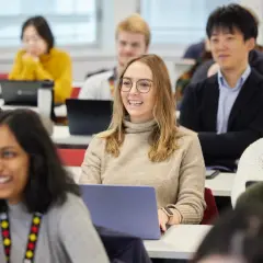 Students in a classroom, some with laptops, are engaged in a lesson. One student smiles, perhaps anticipating her MPA credential.