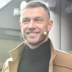 Uśmiechnięty Dariusz Pachut w beżowym płaszczu i czarnym golfie, z mikrofonem przy uchu. Może opowiada o swojej żonie.