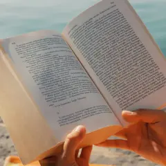 Mani che sfogliano un libro aperto, con un mare azzurro sullo sfondo. Un'immagine che evoca la lettura di una **canzone in dialetto siciliano** sotto il sole.