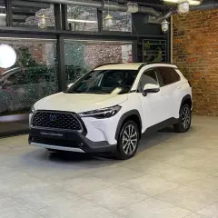 Biała Toyota Corolla Cross Hybrid stoi w nowoczesnym wnętrzu z ceglaną ścianą i dużymi oknami.