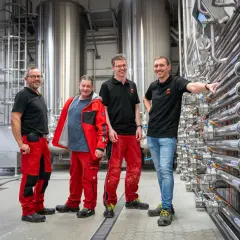 Vier Männer in Arbeitskleidung stehen vor großen Edelstahltanks und Rohrleitungen. Sie sind Teil einer Ausbildung zum Brauer und blicken stolz in die Kamera.