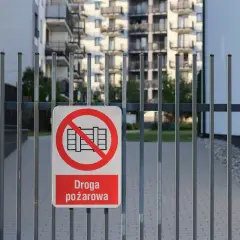 Znak zakazu wjazdu na drogę pożarową na metalowej bramie. W tle budynki mieszkalne i polska flaga.