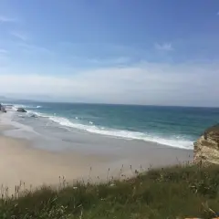 Un paseo solitario por las **playas de Lugo**, con el mar turquesa rompiendo suavemente en la arena dorada y acantilados rocosos.