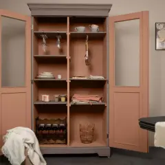 Une vieille armoire repeinte, aux portes roses et intérieur gris, abrite vaisselle, verres à pied, bouteilles de vin et paniers.